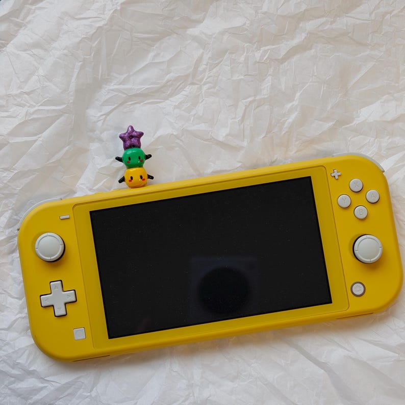 K&ouml;nnte beinhalten: Eine leuchtend gelbe Handheld-Spielkonsole mit schwarzem Bildschirm. Eine kleine, bunte Figur aus gestapelten Charakteren sitzt obenauf. Die Konsole verf&uuml;gt &uuml;ber ein Steuerkreuz, Tasten und Steuerkn&uuml;ppel. Der Hintergrund ist eine strukturierte wei&szlig;e Oberfl&auml;che.
