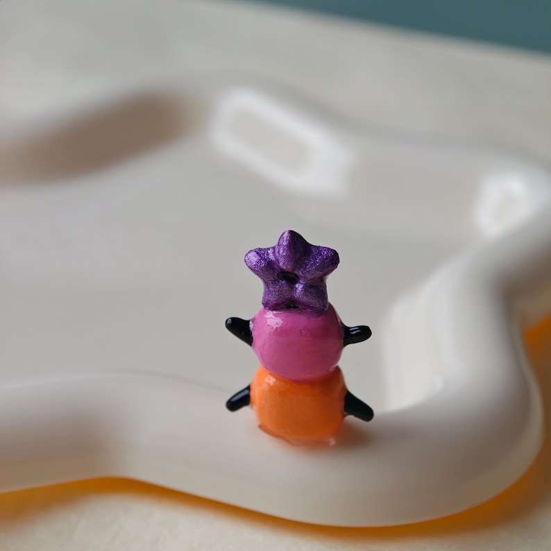 K&ouml;nnte beinhalten: Eine kleine, farbenfrohe Figur einer Person mit einem sternf&ouml;rmigen Hut. Die Figur besteht aus drei &uuml;bereinander gestapelten Kugeln in Pink, Orange und Lila. Sie hat kleine schwarze Arme und steht auf einer wei&szlig;en, geschwungenen Oberfl&auml;che.