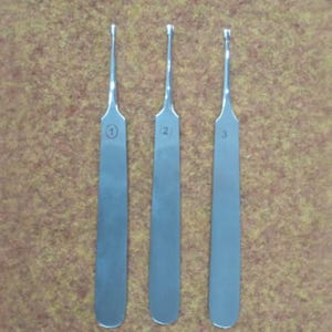 Op de afbeelding: Drie chirurgische instrumenten van roestvrij staal met lange, platte handvatten en genummerde markeringen. Elk gereedschap heeft een dunne, gebogen punt. De achtergrond is een getextureerd, bruin oppervlak.