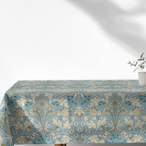 Puede incluir: Un mantel rectangular con un estampado floral en tonos turquesa, beige y dorado. El mantel está sobre una mesa de madera, con una pequeña maceta blanca con vegetación en el fondo. El patrón presenta flores y hojas estilizadas.