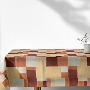 Op de afbeelding: Een rechthoekig tafelkleed met een geometrisch patroon van vierkanten in bruin-, beige- en wittinten. Het tafelkleed is gedrapeerd over een houten tafel, met een kleine plant in een pot op de achtergrond.