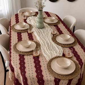May include: An oval dining table covered with a patterned tablecloth featuring vertical stripes in deep red and beige with small blue cross motifs. The table is set with six woven placemats, cream-colored plates, bowls, glasses, and a central vase.