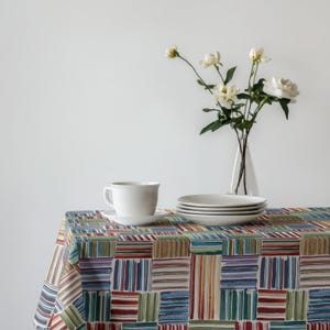 May include: A white ceramic teacup and saucer sit on a colorful tablecloth with a geometric pattern. A stack of white plates and a vase of white flowers complete the table setting. The tablecloth features stripes in various colors.
