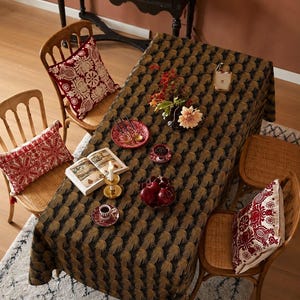Puede incluir: Un mantel rectangular con un patrón repetitivo de abanicos en negro y dorado. La tela cubre una mesa de comedor dispuesta con un plato rojo, un cuenco pequeño con bayas oscuras, un cuenco con manzanas rojas, un libro abierto y un candelabro de latón.