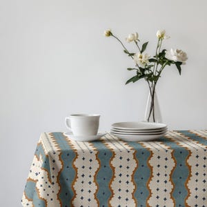 May include: A white ceramic teacup and saucer sit on a patterned tablecloth with a blue, tan, and white design. A stack of white plates and a vase of white flowers complete the table setting.