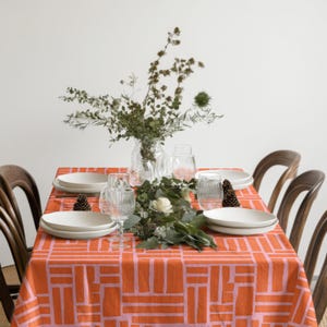 May include: A dining table set for a meal, featuring a vibrant orange and pink patterned tablecloth. White plates, clear glasses, and a floral arrangement with greenery and white flowers adorn the table. Wooden chairs surround the table, creating a warm and inviting atmosphere.