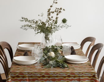 Peut inclure: Une table à manger dressée avec des assiettes blanches, des verres transparents et une composition florale. La table est recouverte d'une nappe à motifs orange, jaune et bleu. Des chaises en bois entourent la table.