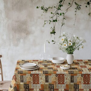 May include: A dining table set with a patterned tablecloth in shades of brown, beige, and red. White plates, a bowl, and a vase with white flowers are on the table. A lit candle and glasses are also present.