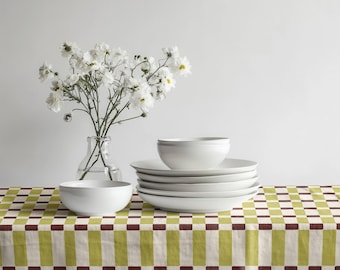 May include: A still-life composition featuring white ceramic dinnerware, including bowls and plates, arranged on a patterned tablecloth. A clear glass vase holds white flowers. The tablecloth has a green, cream, and burgundy checkered design.