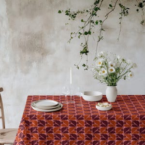 May include: A dining table set with a patterned tablecloth in shades of red and orange. The table is set with white plates, a bowl, and a vase of white flowers. A lit candle and wine glasses are also present.