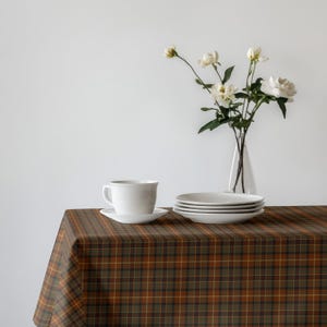 May include: A white teacup and saucer sit on a plaid tablecloth with a brown, orange, and green pattern. A stack of white plates and a vase of white flowers complete the table setting.