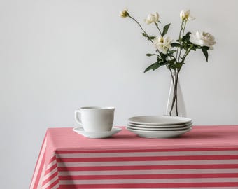 May include: A white teacup and saucer sit on a red and white striped tablecloth, next to a stack of plates and a vase of white flowers. The scene is set against a white background, creating a clean and inviting aesthetic.