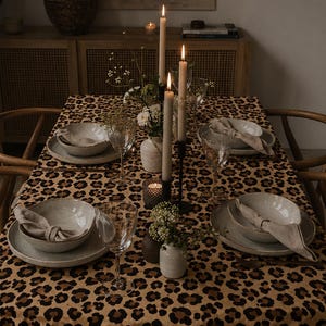 Könnte beinhalten: Ein Esstisch mit einer beigefarbenen Tischdecke mit braun-schwarzem Leopardenmuster. Der Tisch ist mit hellgrauen Keramikschalen und Tellern, beigen Leinenservietten, klaren Weingläsern und drei hohen, brennenden Stabkerzen eingedeckt.