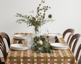 May include: A dining table set for a meal, featuring a tablecloth with a woven pattern in olive green, white, and dusty rose. White plates, clear glasses, and a floral arrangement complete the setting. Wooden chairs surround the table.
