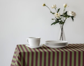 May include: A white teacup and saucer sit on a striped tablecloth with vertical burgundy and olive green stripes. White plates are stacked nearby, and a vase of white flowers adds a touch of elegance to the scene.