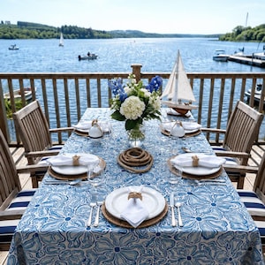 May include: A dining table set for four on a wooden deck overlooking a lake, featuring a blue and white floral tablecloth, white plates with rope napkin rings, a central vase of white and blue flowers, and a small decorative wooden sailboat model.