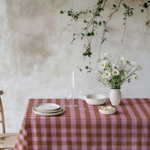 May include: A dining table set with a pink and brown gingham tablecloth. The table is set with white plates, a bowl, and a vase of white flowers. A lit candle and two wine glasses are also on the table.