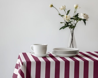 May include: A white teacup and saucer sit on a striped burgundy and white tablecloth. A stack of white plates and a vase of white flowers complete the table setting. The tablecloth has vertical stripes.