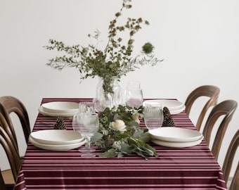 May include: A dining table set for a meal, featuring a burgundy tablecloth with pink stripes. White plates, clear glasses, and a floral arrangement with greenery and white flowers are arranged on the table. Wooden chairs surround the table.