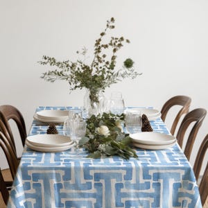 May include: A dining table set with a blue and white patterned tablecloth. White plates, clear glasses, and a floral centerpiece adorn the table. Wooden chairs surround the table, creating a welcoming atmosphere for a meal.