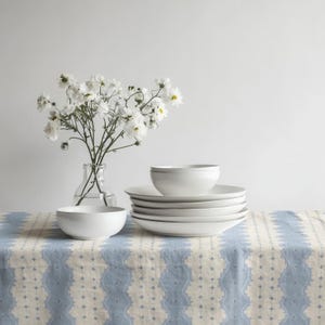 Puede incluir: Un bodegón con vajilla de cerámica blanca, que incluye cuencos y platos, colocada sobre un mantel con estampado azul y blanco. Un jarrón de cristal transparente contiene flores blancas, añadiendo un toque natural.