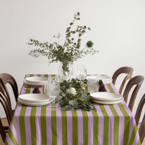 May include: A dining table set with a green and lilac striped tablecloth. White plates, clear glasses, and a floral arrangement with greenery and pine cones are arranged on the table. Wooden chairs surround the table setting.
