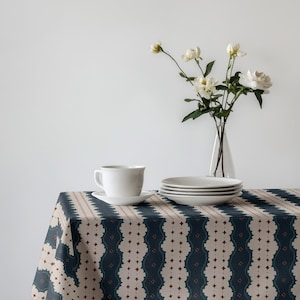 May include: A table setting featuring a white teacup and saucer, stacked plates, and a vase of white flowers. The table is covered with a patterned tablecloth in navy blue, beige, and burgundy.