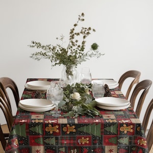 Op de afbeelding: Een feestelijke tafelsetting met een kerstkleed met peperkoekenmannetjes en kerstbomen. Witte borden, heldere glazen en een bloemstuk. Houten stoelen omringen de tafel.