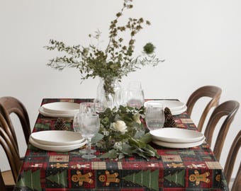 May include: A festive table setting with a Christmas-themed tablecloth featuring gingerbread men and Christmas trees. White plates, clear glasses, and a floral centerpiece complete the holiday decor. Wooden chairs surround the table.