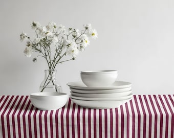 May include: A still-life image of white ceramic dinnerware, including bowls and plates, arranged on a striped burgundy and white tablecloth. A clear glass vase with white flowers is also present, creating a minimalist aesthetic.