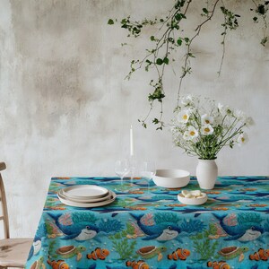 May include: A blue tablecloth with an underwater scene featuring sharks, turtles, and clownfish. The table is set with white plates, a bowl, and a vase of white flowers. A candle and wine glasses are also on the table.