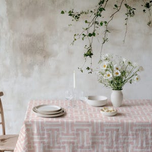 May include: A dining table set with a pink and white geometric patterned tablecloth. The table is set with white plates, a white bowl, and a vase of white flowers. A lit candle and two wine glasses are also on the table.