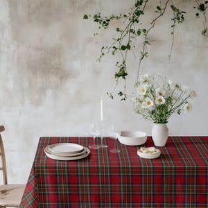 May include: A red and green plaid tablecloth covers a table set for a meal. White plates, a bowl, and a vase of white flowers are on the table. Two wine glasses and a candle are also present, creating a festive dining scene.