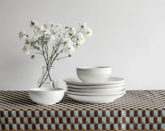 May include: A still life arrangement featuring white ceramic dinnerware, including bowls and plates, stacked on a patterned tablecloth. A clear glass vase holds white flowers, adding a touch of nature to the scene.