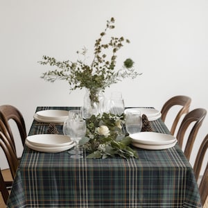 May include: A dining table set for a meal, featuring a green and blue plaid tablecloth. White plates, clear glasses, and a floral centerpiece with greenery and white flowers are arranged on the table. Wooden chairs surround the table.