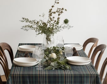 May include: A dining table set for a meal, featuring a green and blue plaid tablecloth. White plates, clear glasses, and a floral centerpiece with greenery and white flowers are arranged on the table. Wooden chairs surround the table.