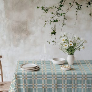 May include: A dining table set for a meal, featuring a blue and beige checkered tablecloth. The table is set with white plates, a bowl, and a vase of white flowers. A lit candle and glasses are also present.