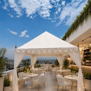 May include: A white square canopy tent with scalloped edges and draped curtains stands on a rooftop terrace. The area features multiple round tables with white chairs, potted plants, and a bar counter under a partially covered section.