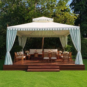 May include: A large outdoor pavilion featuring a white canopy with a scalloped edge and green and white striped curtains tied at the corners. The structure sits on a raised wooden deck furnished with wooden chairs and sofas with light cushions.