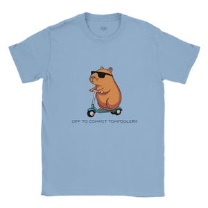 Op de afbeelding: Lichtblauw T-shirt met een cartoon capibara met een zonnebril en een turquoise scooter. De tekst "OFF TO COMMIT TOMFOOLERY" staat eronder. Het shirt is gemaakt van zacht materiaal.