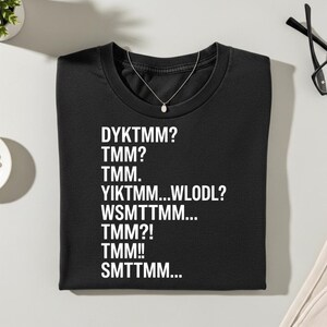 May include: Black t-shirt with white text: DYKTMM?, TMM?, TMM., YIKTMM...WLODL?, WSMTTMM..., TMM?!, TMM!!, SMTTMM... A silver necklace with a small pendant is on the shirt. The shirt is folded neatly on a white surface.