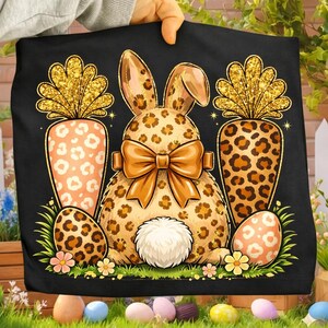 May include: Black Easter-themed graphic featuring a leopard print bunny with a large bow, two carrots, and two decorated eggs. The carrots have gold glitter tops. The bunny has a white fluffy tail. The design is set against a black background.