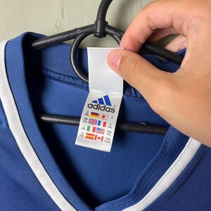 May include: A blue V-neck shirt with white trim around the collar. A white interior tag features the Adidas logo in blue and lists size 2XL for multiple countries including Germany, Japan, Great Britain, France, Spain, USA, and Canada with flags.