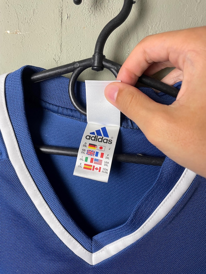 May include: A blue V-neck shirt with white trim around the collar. A white interior tag features the Adidas logo in blue and lists size 2XL for multiple countries including Germany, Japan, Great Britain, France, Spain, USA, and Canada with flags.