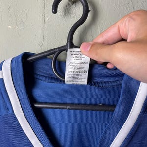 May include: A blue athletic shirt with white trim along the neckline, hanging on a black plastic hanger. A white care label is attached to the collar, featuring text in multiple languages including '100% Polyester' and washing instructions.