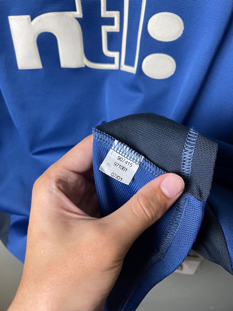 May include: A blue athletic jersey featuring the white text ntl: printed on the front. An interior white fabric tag is visible, displaying the numbers 907415, 971001, and the date 07/01. The garment is constructed from a textured, breathable material.