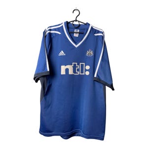 May include: A blue short-sleeved soccer jersey with a V-neck collar, white trim, and white stripes on the shoulders. The shirt features an Adidas logo, the Newcastle United crest, and the text ntl: printed in white on the front chest area.