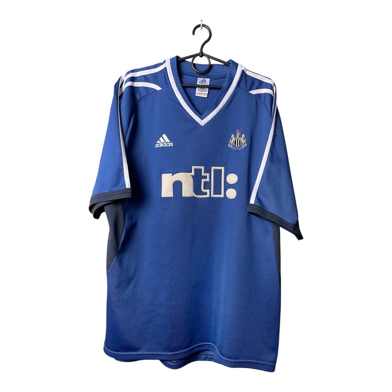 May include: A blue short-sleeved soccer jersey with a V-neck collar, white trim, and white stripes on the shoulders. The shirt features an Adidas logo, the Newcastle United crest, and the text ntl: printed in white on the front chest area.