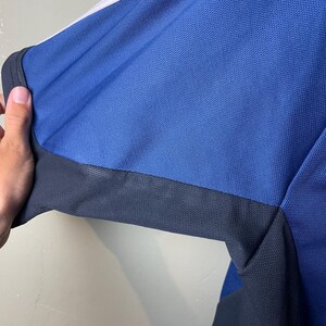 May include: A short-sleeved athletic shirt featuring a bright blue textured mesh fabric. The garment includes a dark charcoal gray panel along the underarm and side, complemented by a crisp white stripe running along the top of the shoulder.
