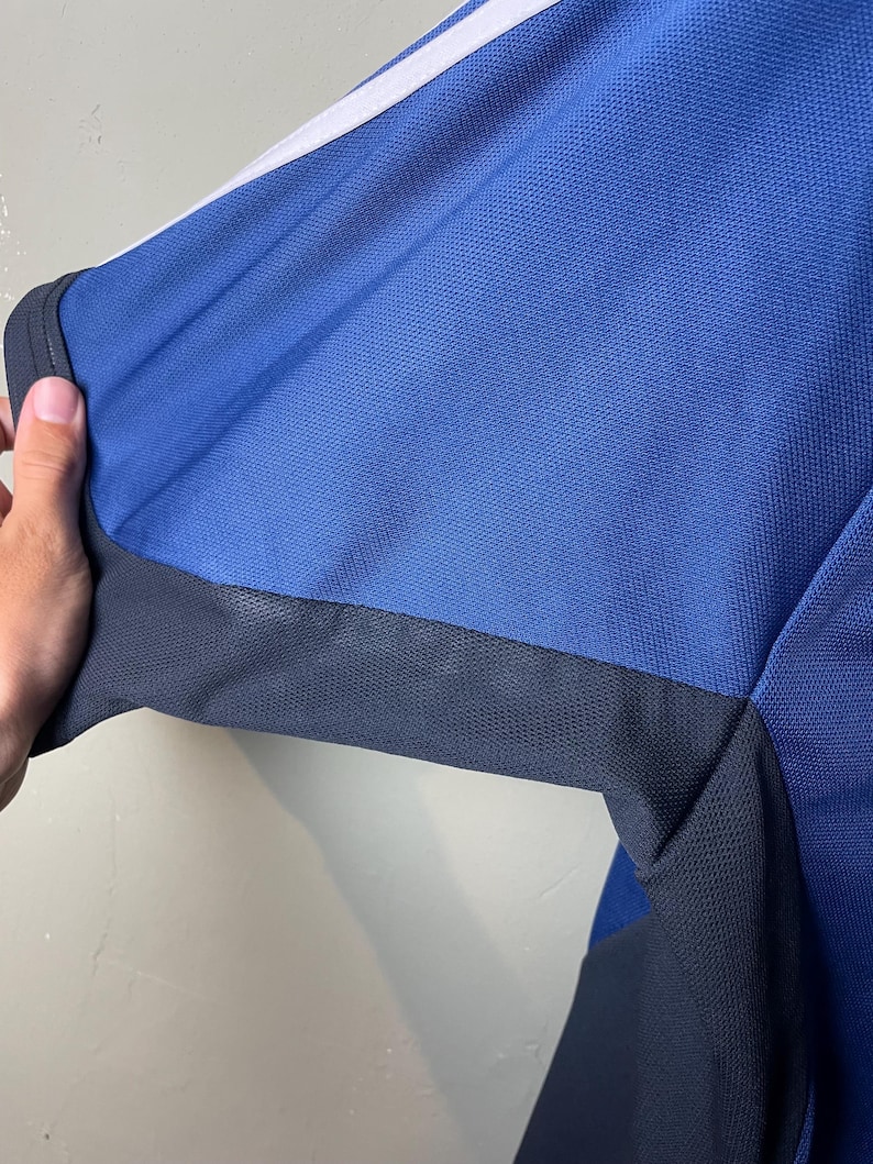 May include: A short-sleeved athletic shirt featuring a bright blue textured mesh fabric. The garment includes a dark charcoal gray panel along the underarm and side, complemented by a crisp white stripe running along the top of the shoulder.
